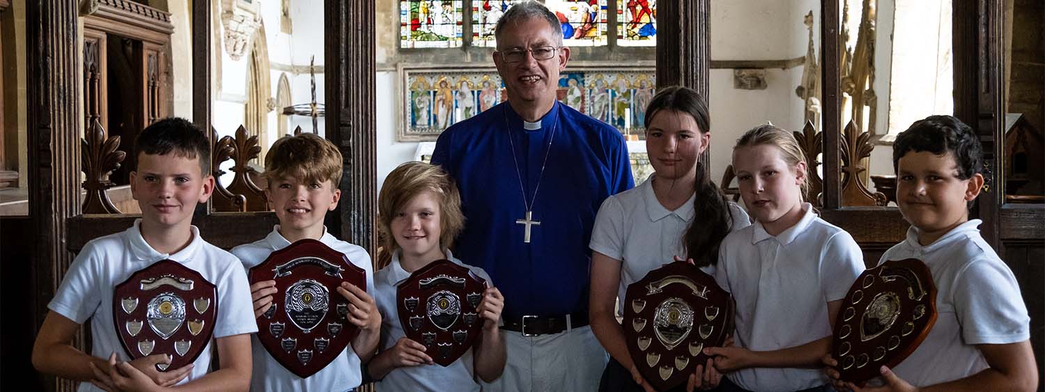 Bishop Steven and Year 6 pupils from Gt Horwood CE School stand in St James' Church, Gt Horwood. The pupils hold shields they have been awarded for excellence in their last year of primary school.