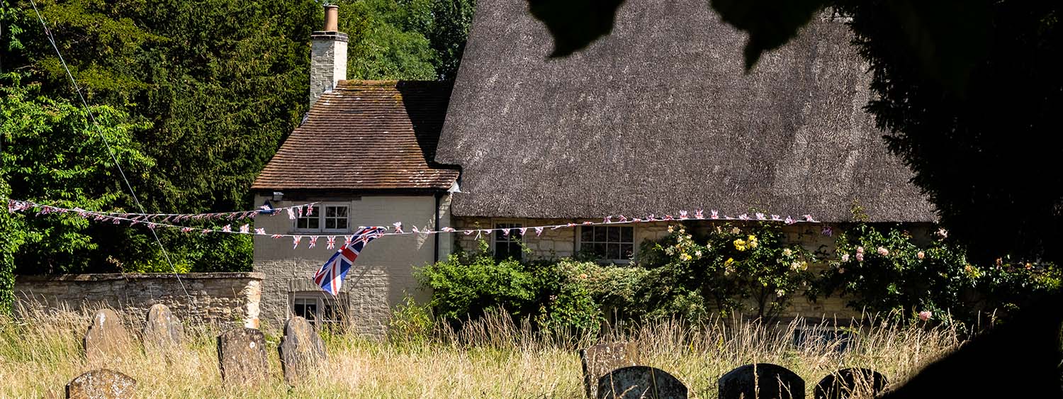 A churchyard backs onto a thatched cottage with Union Jack bunting blowing in the wind