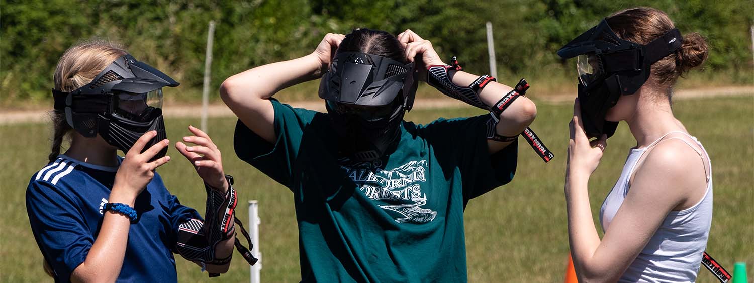 Three teenage girls prepare for a game of archery tag, pulling on black face visors.