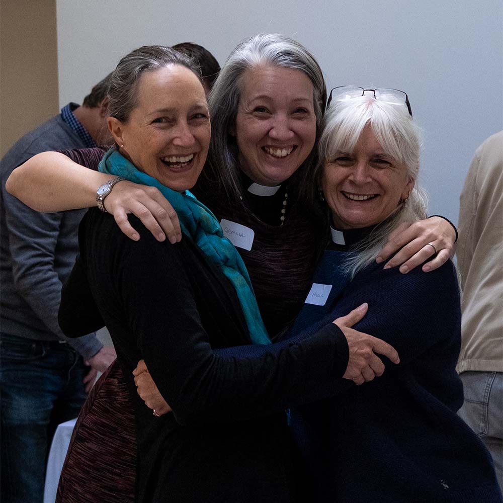 Three women clergy members hug each other and smile for a photo.