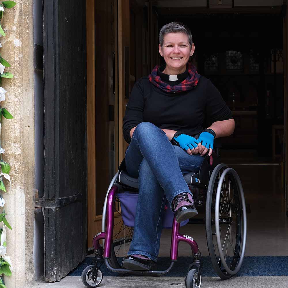 The Revd Katie Tupling sits in her purple wheelchair in the doorway to a church building.