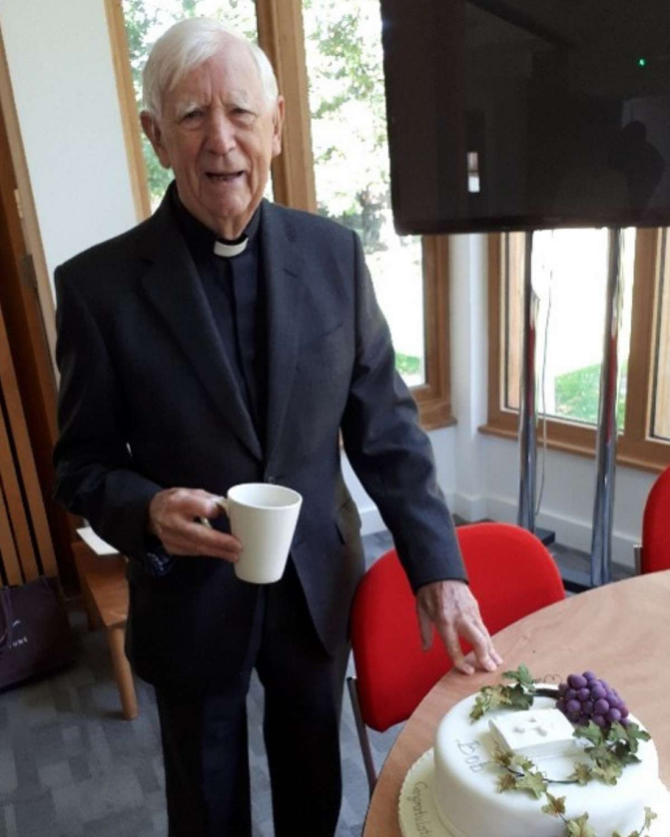 Bob Whiteley standing next a table with a celebration cake on it and he is holding a white coffee cup.