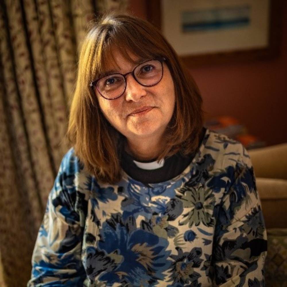 The Reverend Jane Willis is wearing a floral jumper and clerical collar, she is sitting inside infront of a curtain and there is a framed painting on the wall behind her.