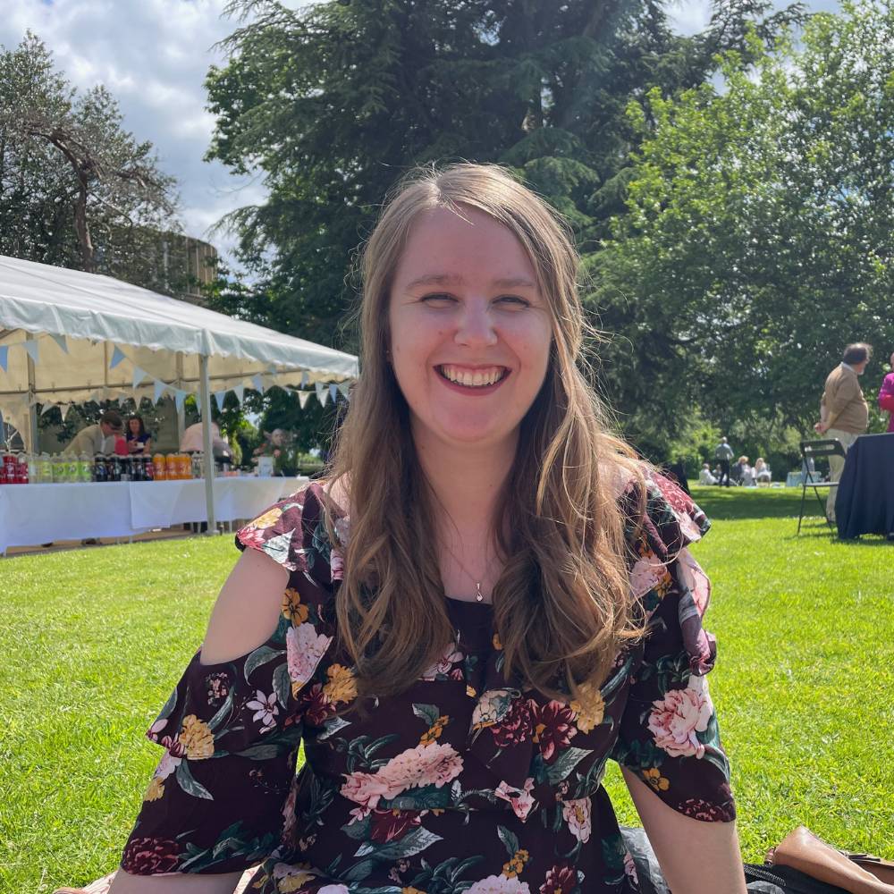 Rebecca sits outside on grass in a garden, she is smiling and there is a marquee in the background.