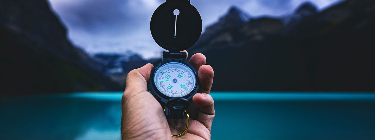 A hand holds a compass aloft as they look out onto a lake