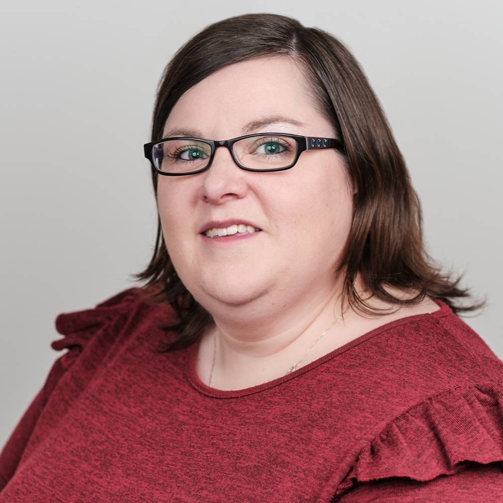 Headshot of Laura-Anne standing side on to the camera. Laura-Anne is smiling, she has brown shoulder length hair and its wearing dark-rimmed glasses.