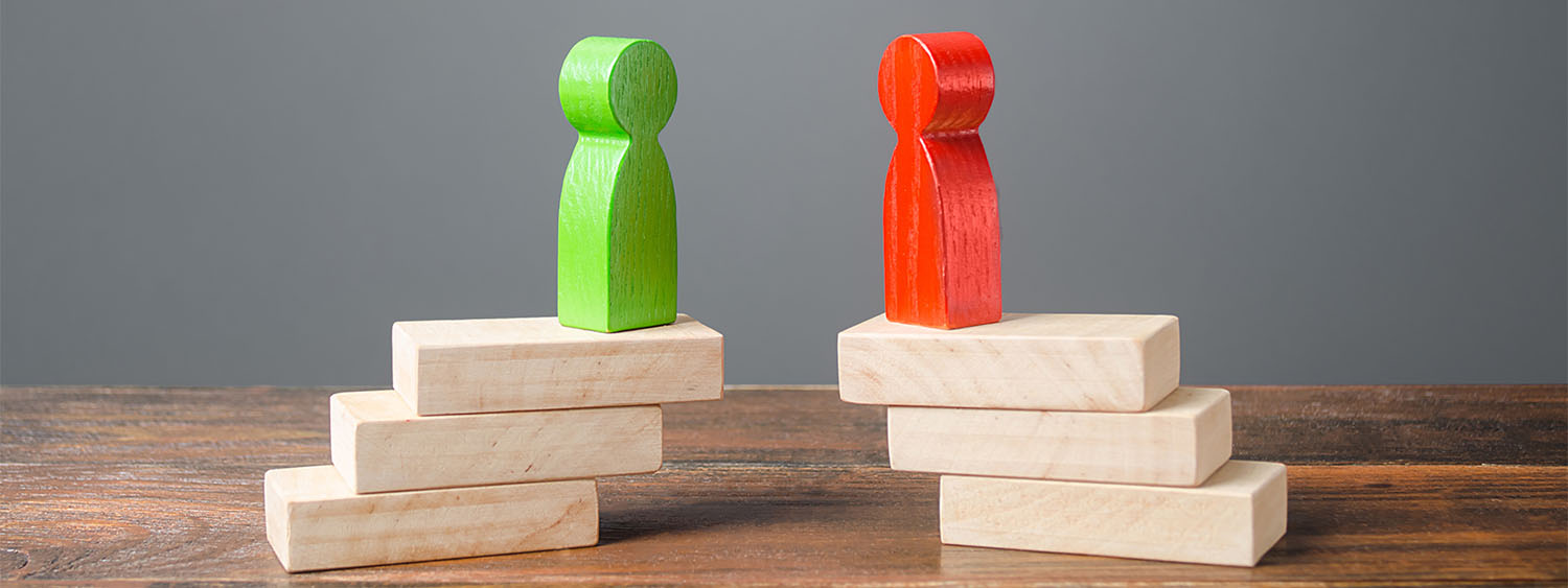 Two wooden figures stand facing each other. Each stands on a small tower of wooden blocks. They can't quite reach each other.