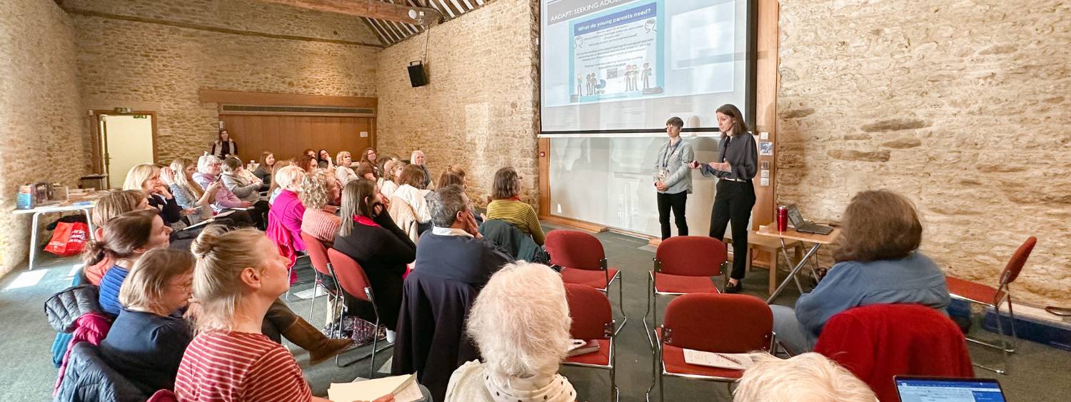 Group of people sit in rows of chairs facing two speakers stood giving a presentation