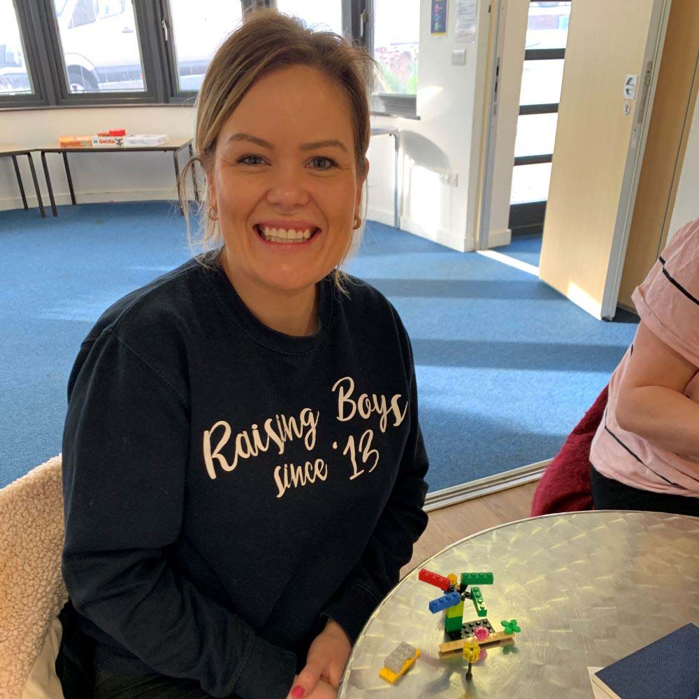 A woman smiles at the camera. A Lego creation is on the table in front of her