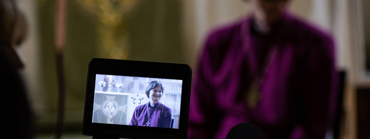 Bishop Olivia is seen on a film camera screen sitting in St Mary's Church wearing a purple cassock and smiling.