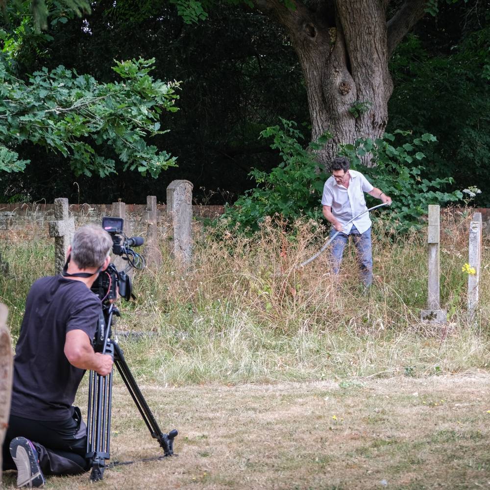 Mike Buckland is scything long grass in the St Mary's churchyard, a man is kneeling with a large camera filming.