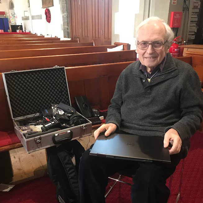 John Trett, a professional hearing loop engineer, sits in a church building with his laptop and sound equipment, smiling at the camera