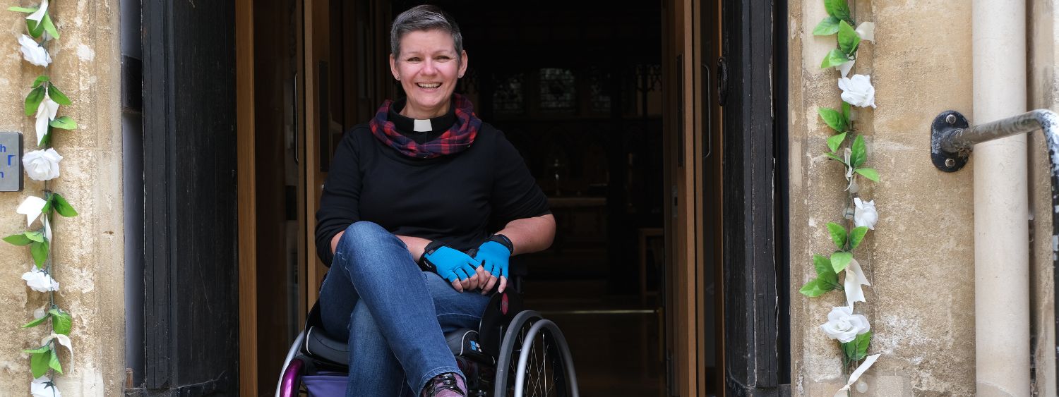 Katie Tupling sits in a wheelchair in church entrance and she is smiling. There is a flower garland draped around the doorway and railings are the church entrance.
