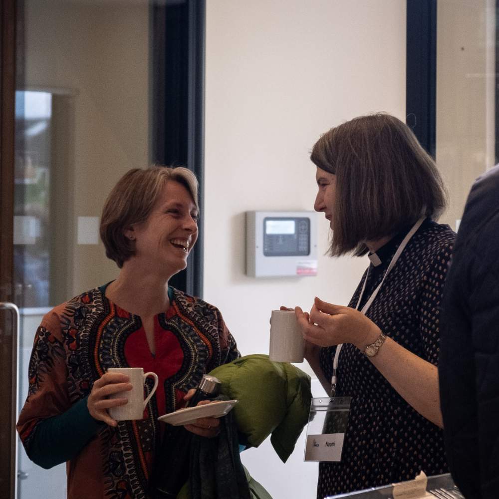 Two attendees stand talking to one another smiling and holding cups of coffee.