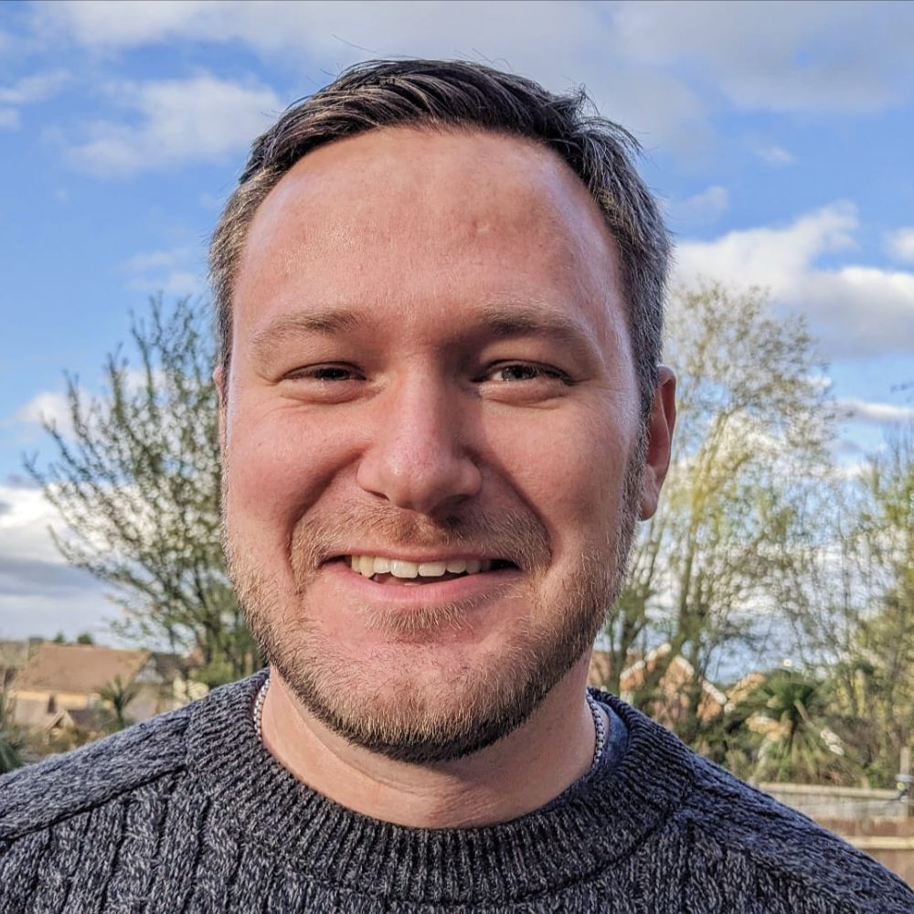 Headshot of Adam smiling, he is wearing a dark grey round neck jumper