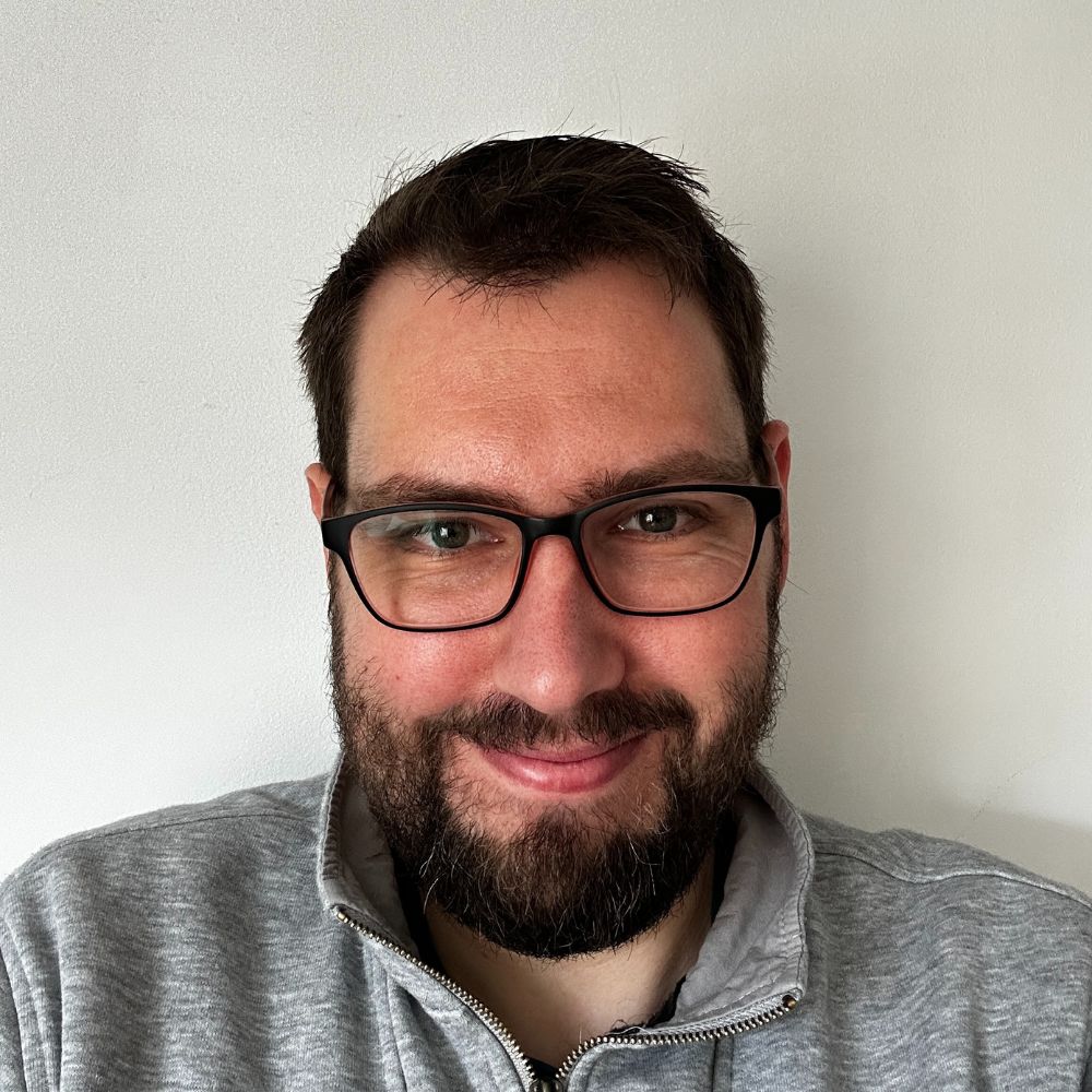 headshot of James, he is smiling and has a beard and is wearing a grey top.