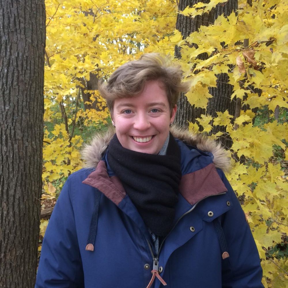 naomi is stood infront of a tree with bright yellow leaves, she is smiling and wearing an navy coat.