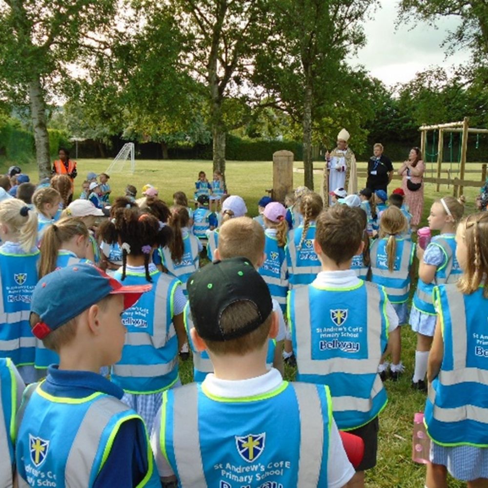 A large group of primary school children in blue hi-vis jackets are standing in a grass with teachers and the Bishop of Dorchester.