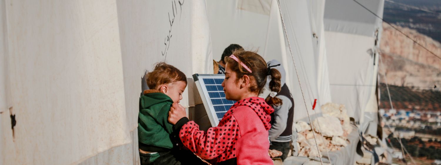 Young girl wearing pink jacket plays with a toddler wearing a green jacket next to tents in a refugee camp.