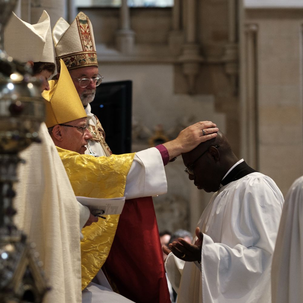 Sydney Quartey kneels before the Bishop of Oxford with his hands raised palms up and the bishop places his hands on his head.